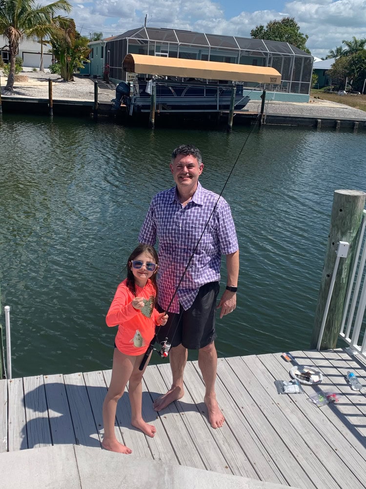 Man and girl fishing on a dock by the water with a boat lift in background.