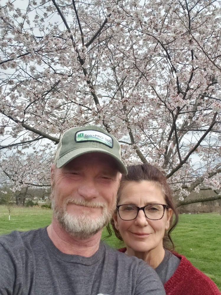 Two people smiling outdoors under blooming cherry blossom trees.
